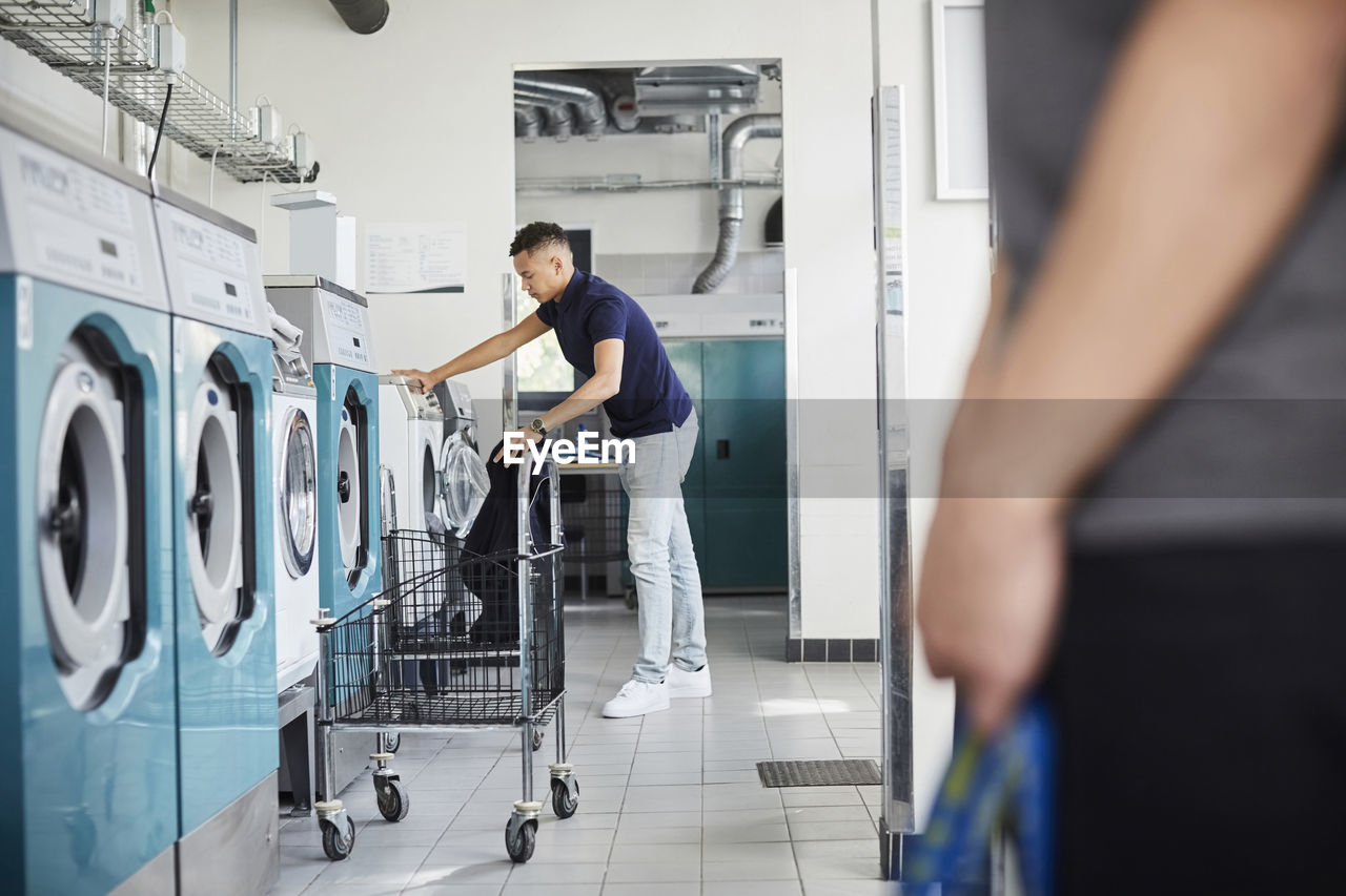 Full length of man using washing machine at laundromat with friend standing in foreground