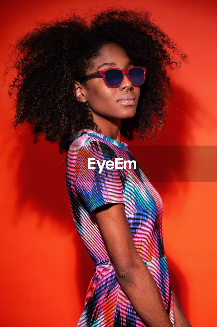 Modern cuban woman with afro hair wearing bright dress and sunglasses in red studio background