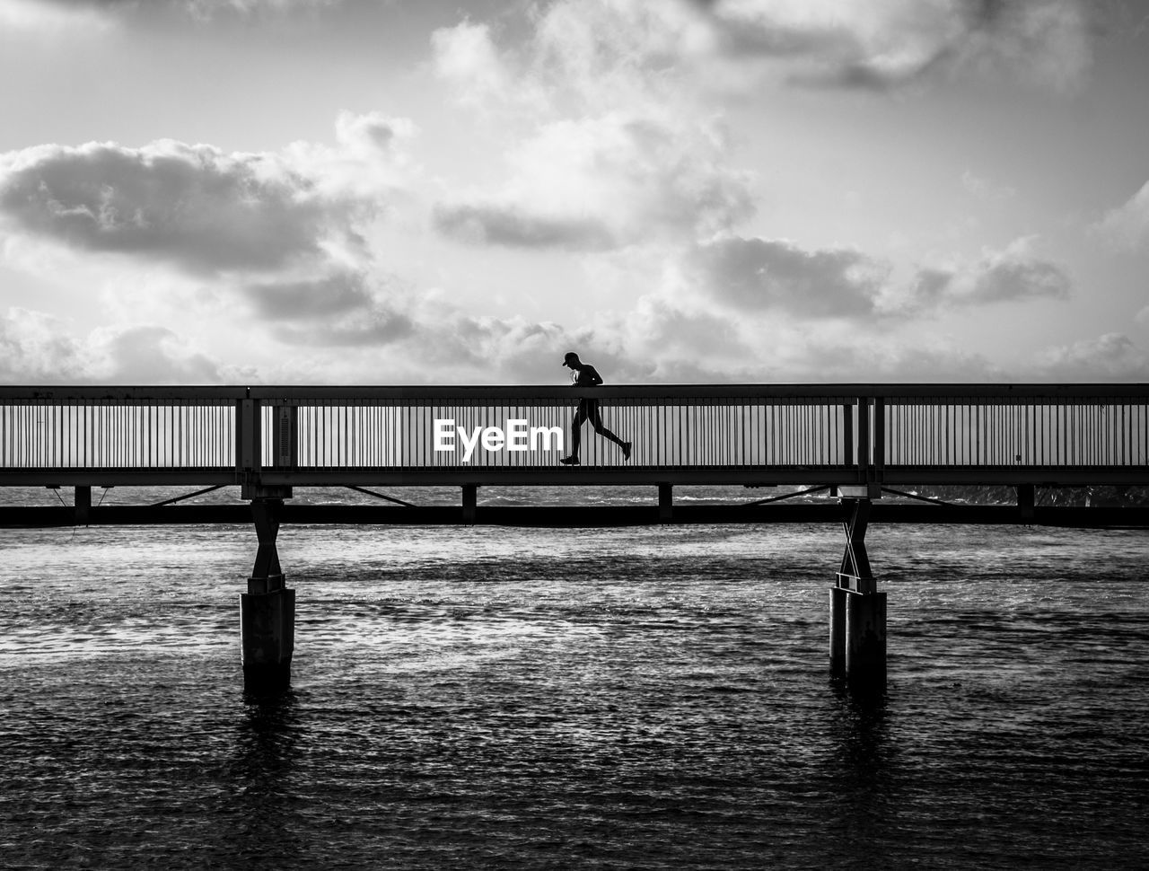 Silhouette man running on bridge over sea against cloudy sky