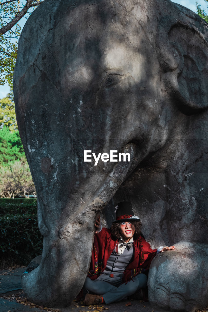 Portrait of man cosplaying mad hatter sitting at the foot of elephant statue