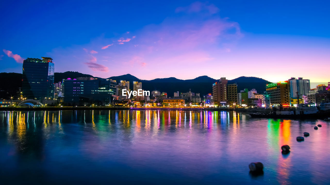 ILLUMINATED BUILDINGS BY LAKE AGAINST SKY AT NIGHT