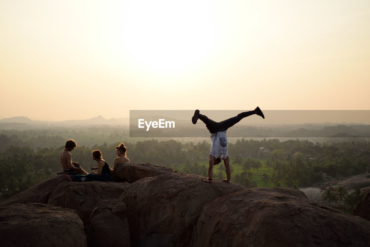 Man doing handstand by friends on rock against sky