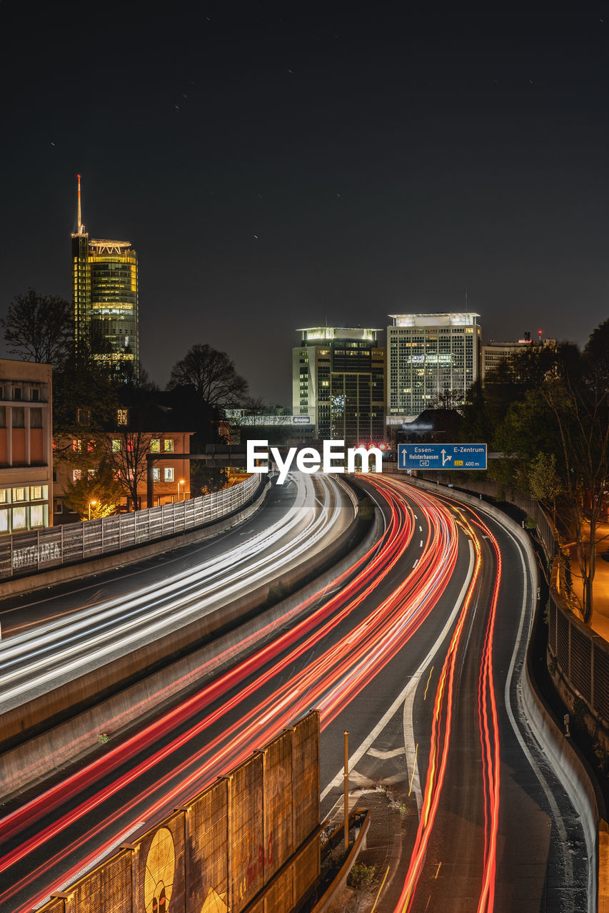 Light trails on road amidst buildings against sky at night