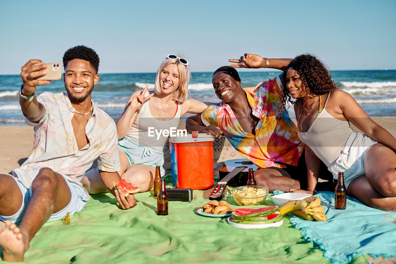 Group of cheerful young multiracial male and female friends in summer clothes smiling and showing two fingers gesture while taking selfie on smartphone during picnic on sandy beach