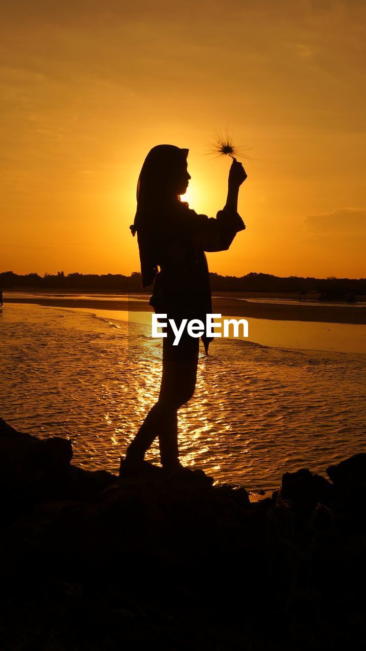 Silhouette girl standing hold dried flower on beach against sky during sunset