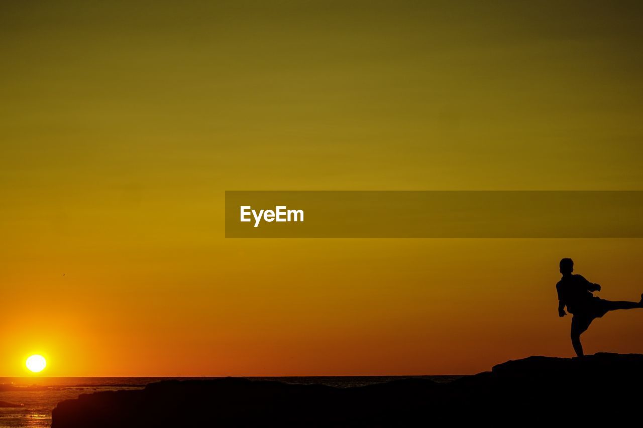 SILHOUETTE MAN STANDING ON ROCK AGAINST ORANGE SKY