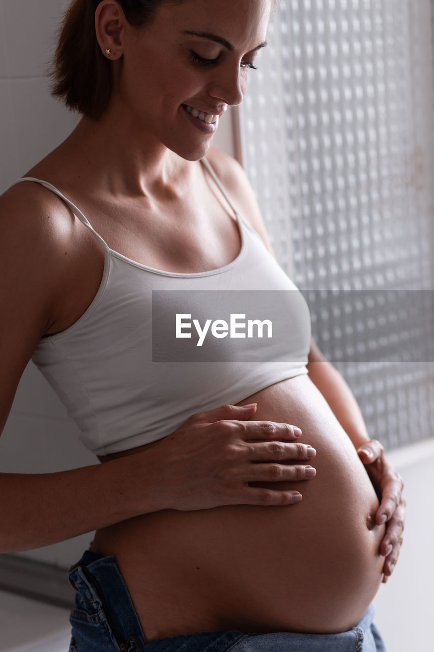 Side view of charming pregnant female smiling and gently touching tummy while standing in room at home