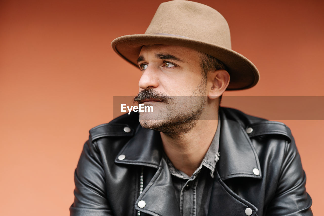 Trendy thoughtful male in hat and leather jacket on beige background looking away