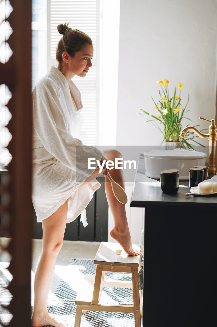 Young woman in white bathrobe doing body massage with dry wooden brush in bathroom at home