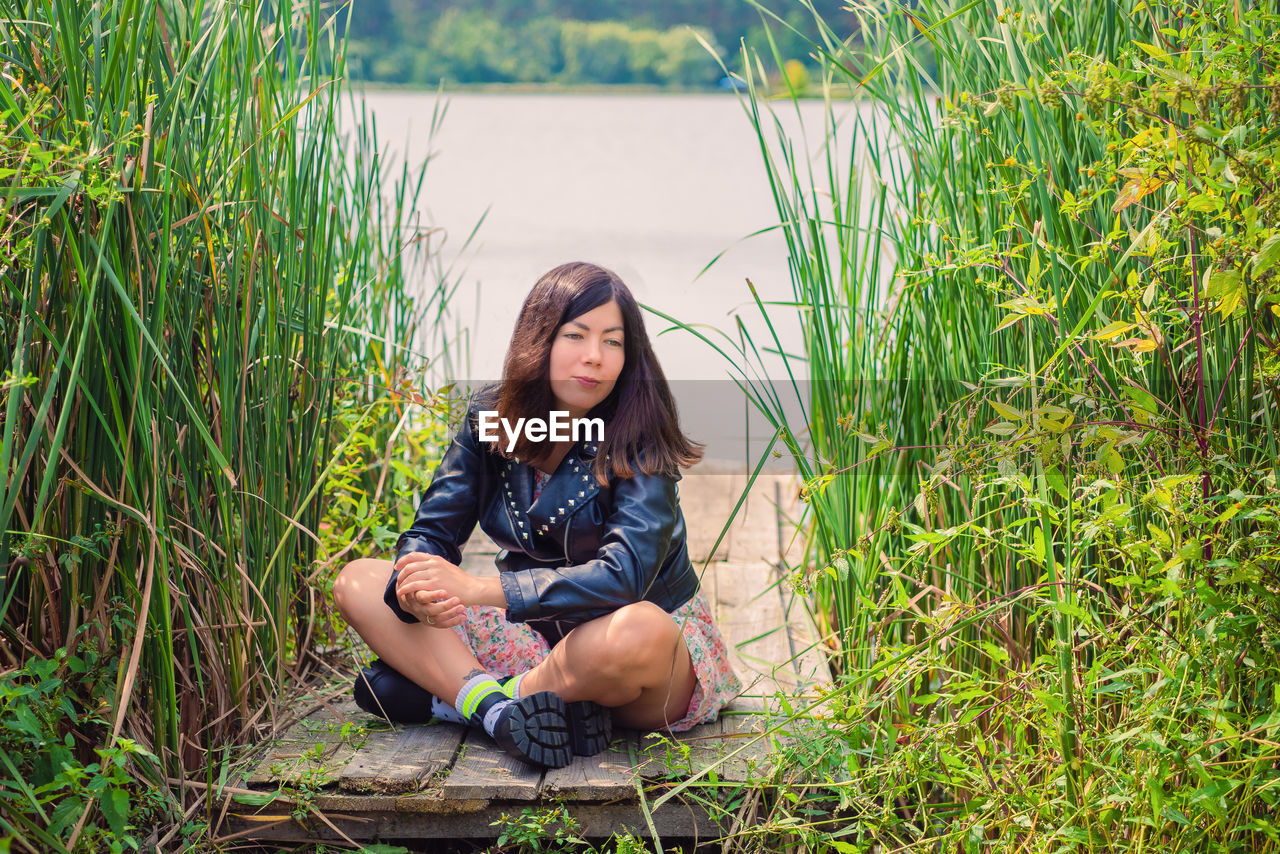 A lovely young woman with black hair is sitting in a thicket of tall grass on the river bank.