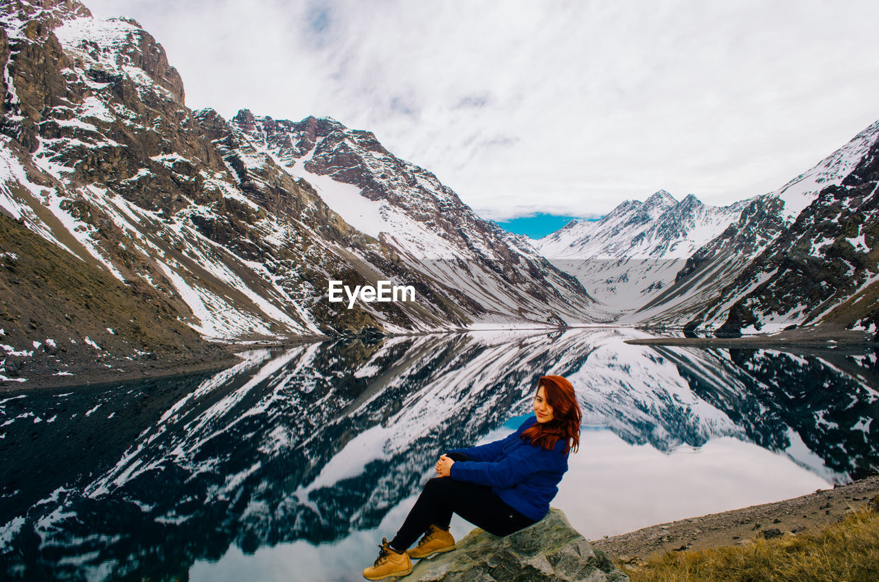 Full length of woman sitting by lake against snowcapped mountain