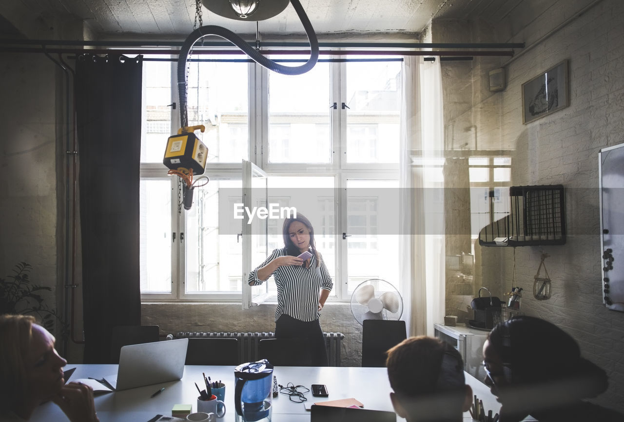 Businesswoman looking at mobile phone while standing against windows in board room