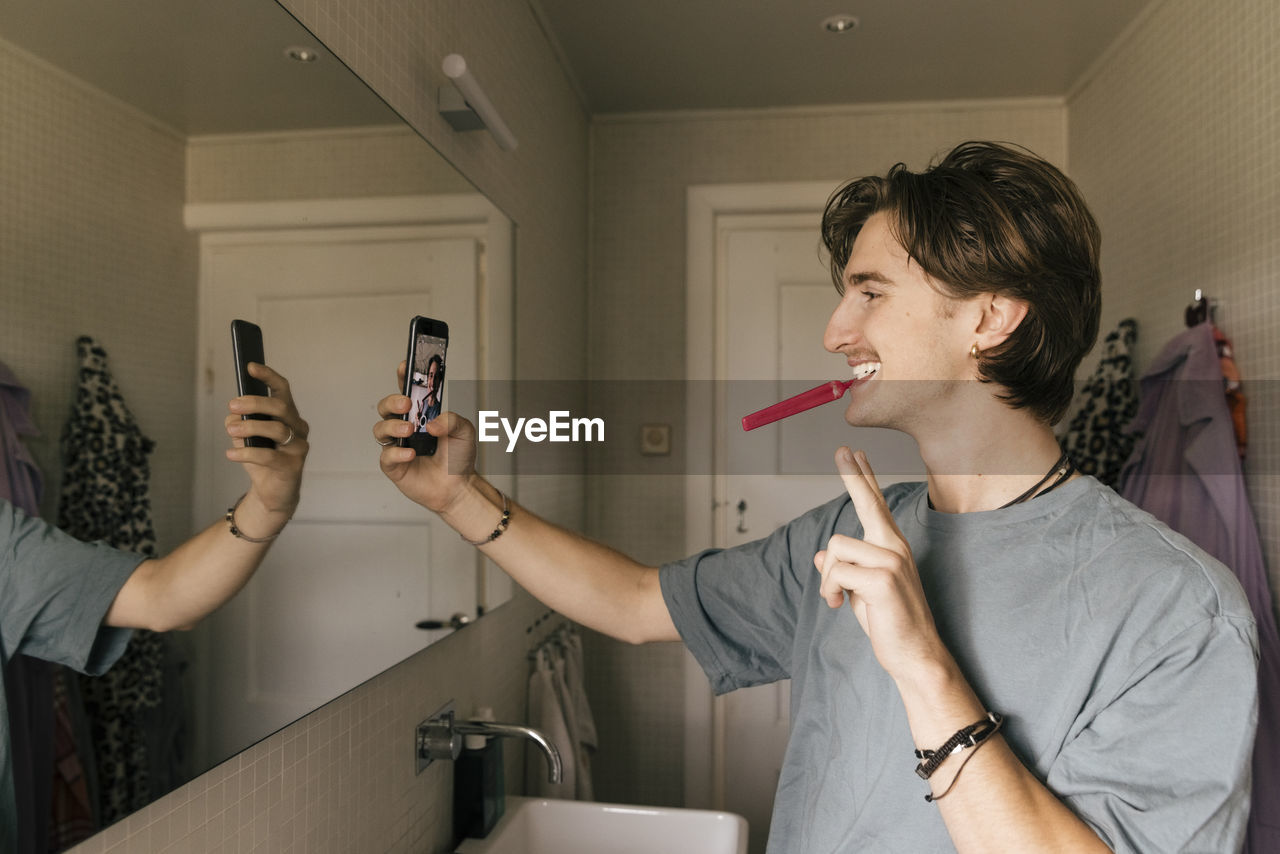 Smiling man taking selfie through smart phone while brushing teeth in bathroom at home