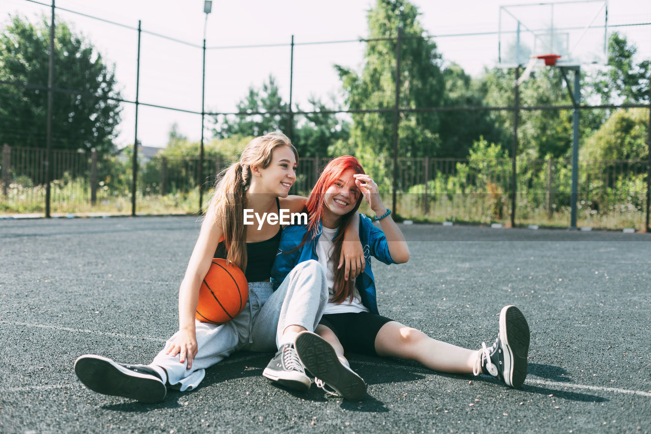 Two girls in sports clothes and with a basketball are chatting, sitting on the playground. 