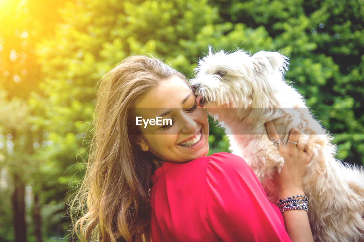 Close-up of dog licking happy woman face against trees in park