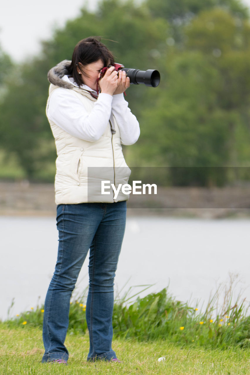 Woman photographing while standing by river on grassy field