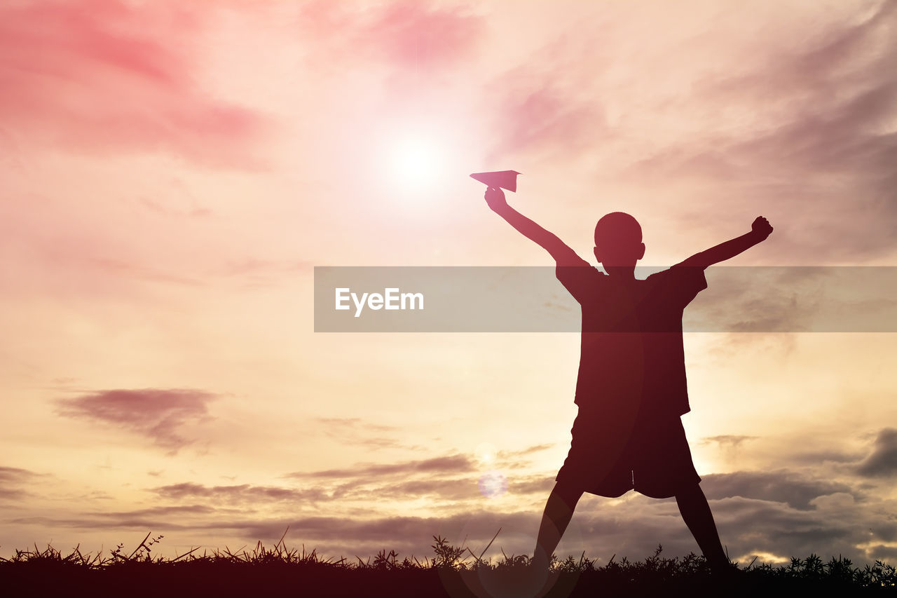 Silhouette boy with paper airplane standing with arms outstretched during sunset