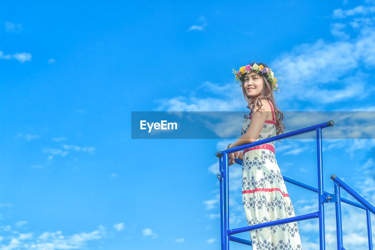 Low angle view of woman wearing flowers while standing by railing against sky