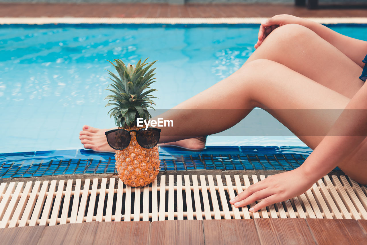 Low section of woman relaxing by pineapple with sunglasses at poolside