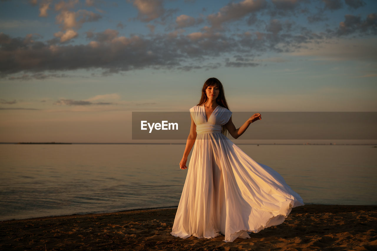 Beautiful young woman in a white long dress near the sea at sunset stands