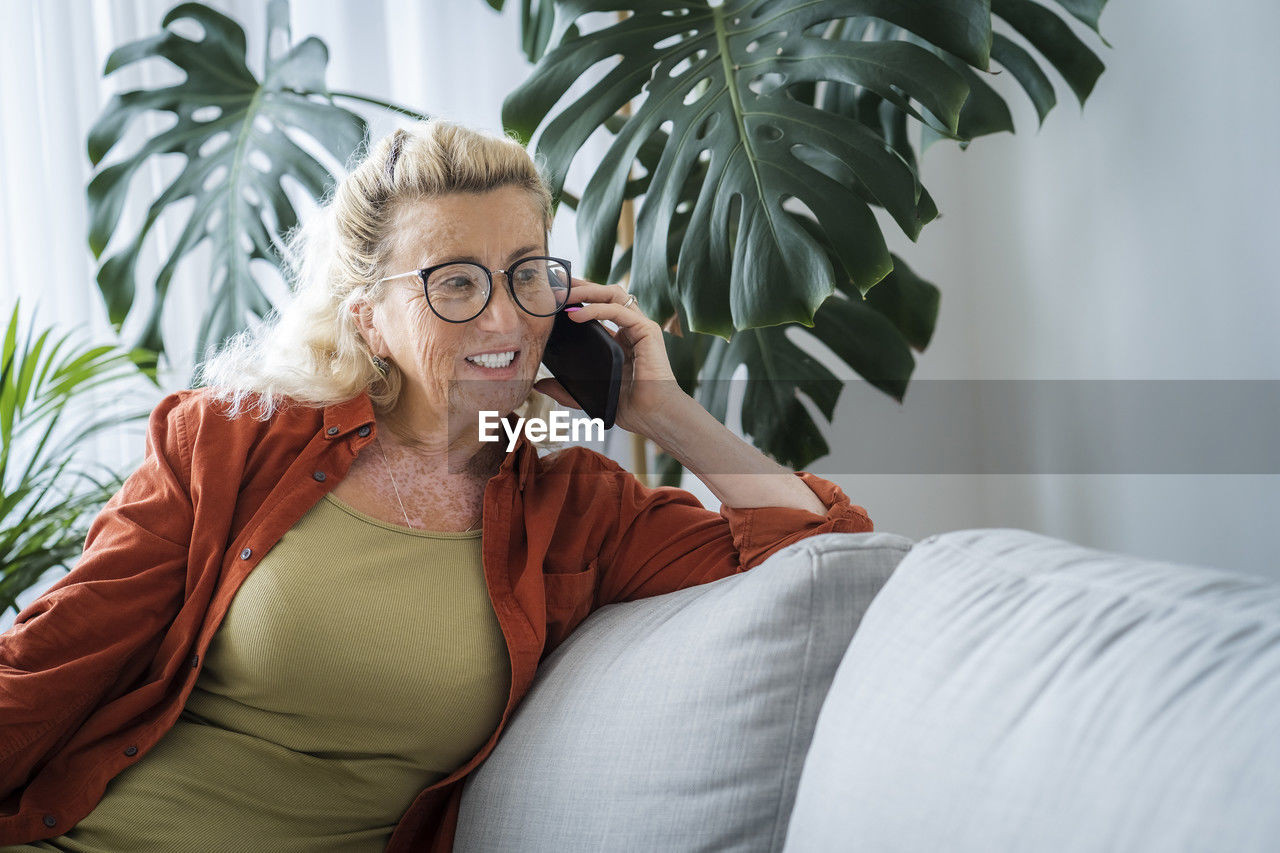 Happy senior woman talking through smart phone leaning on sofa at home