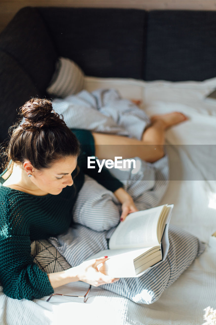 High angle view of young woman reading book while lying on bed at home