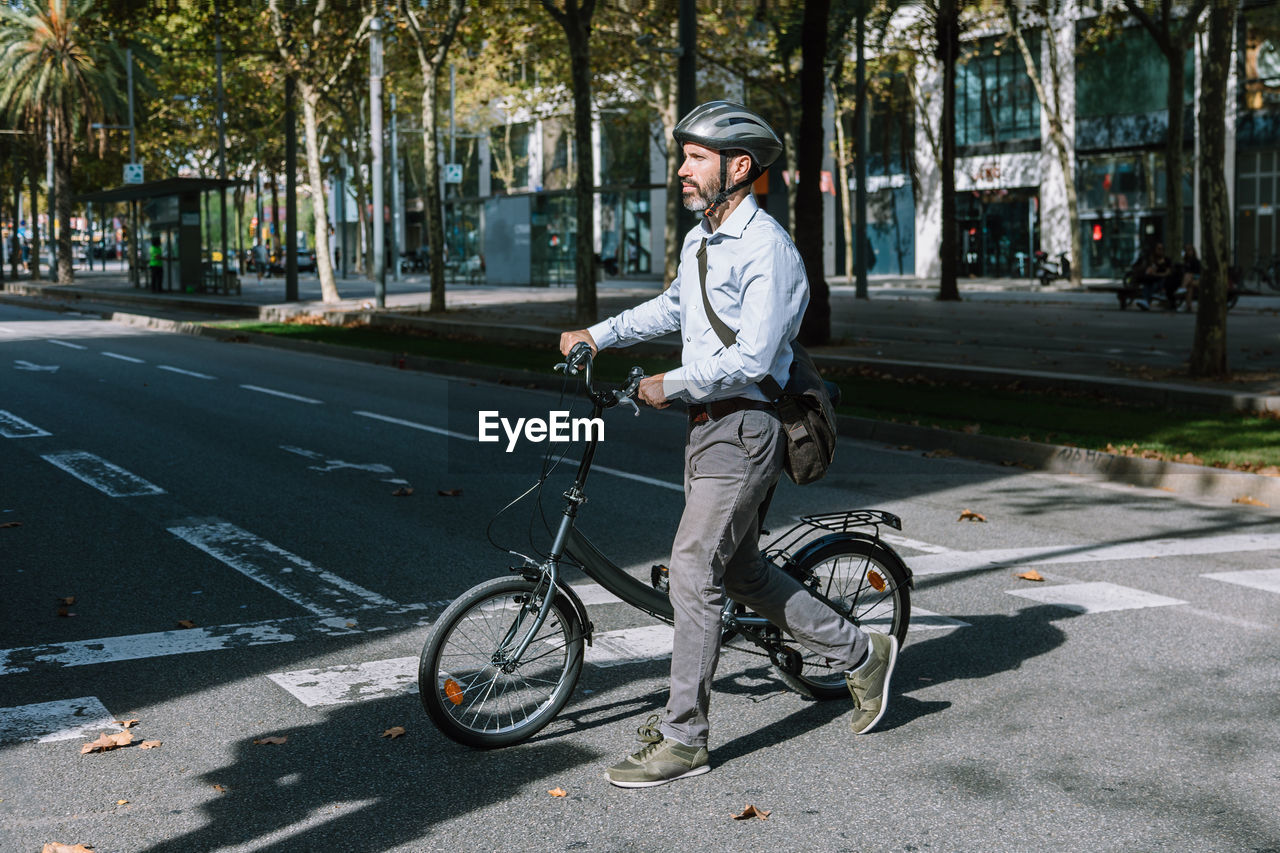 Full length of bearded male worker in helmet crossing asphalt roadway with bicycle on way to work