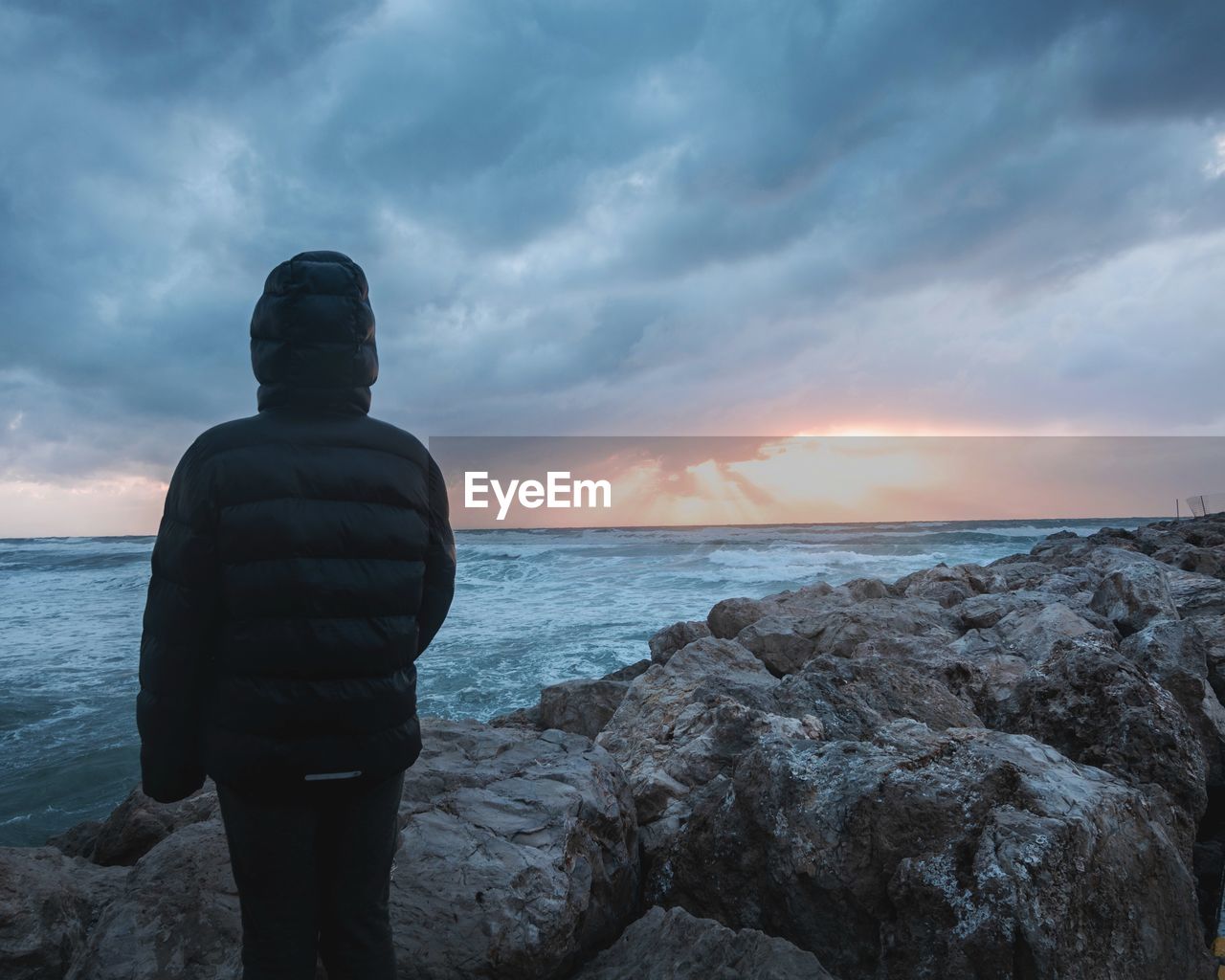 REAR VIEW OF MAN STANDING ON ROCK AT SEA SHORE AGAINST SKY