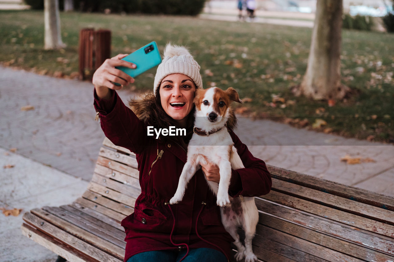 Woman with dog using phone while sitting on bench against trees