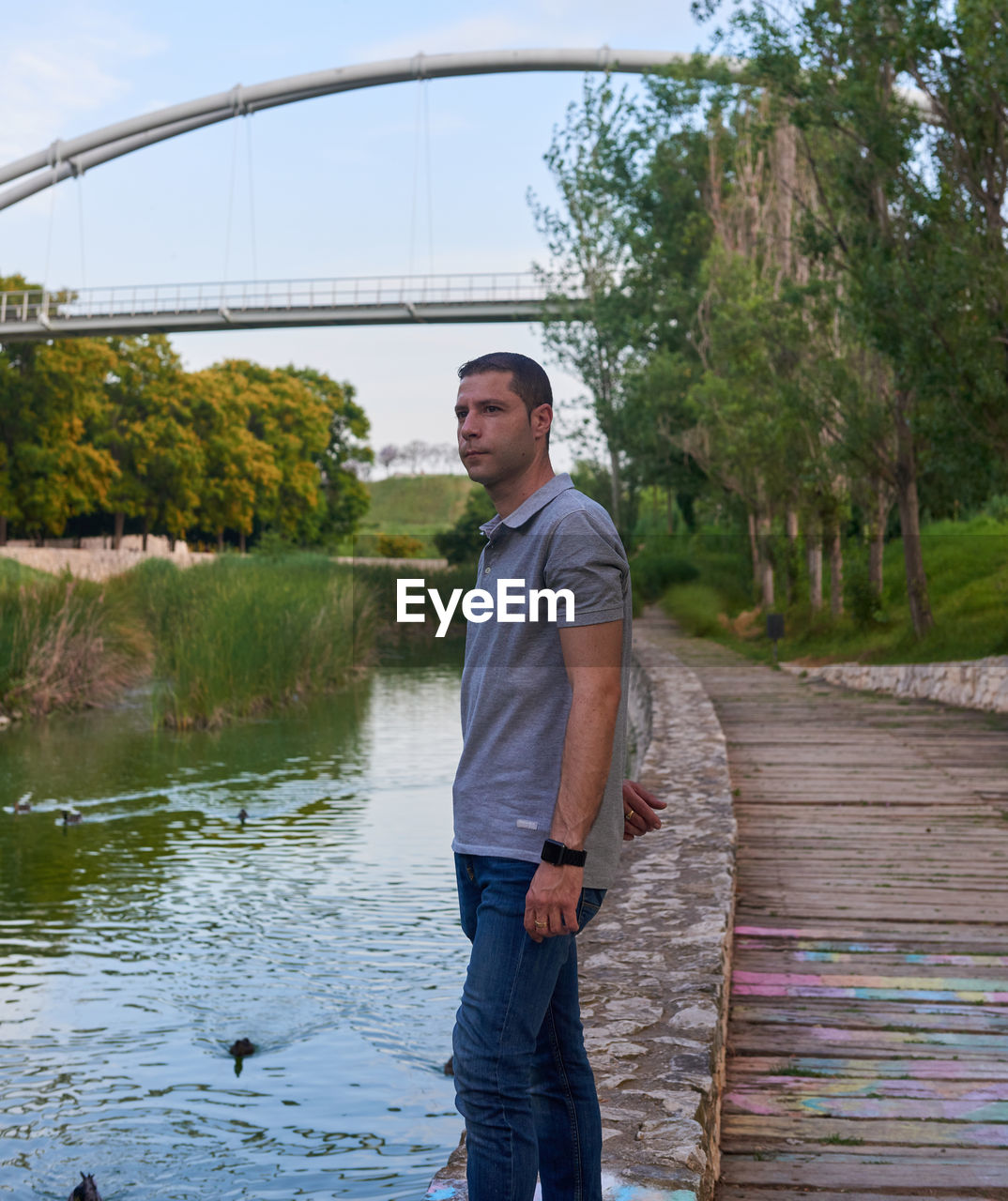 one person, water, adult, men, standing, casual clothing, nature, day, plant, tree, leisure activity, lifestyles, three quarter length, young adult, full length, river, architecture, side view, outdoors, looking, sky, bridge, animal, bird, smiling, jeans