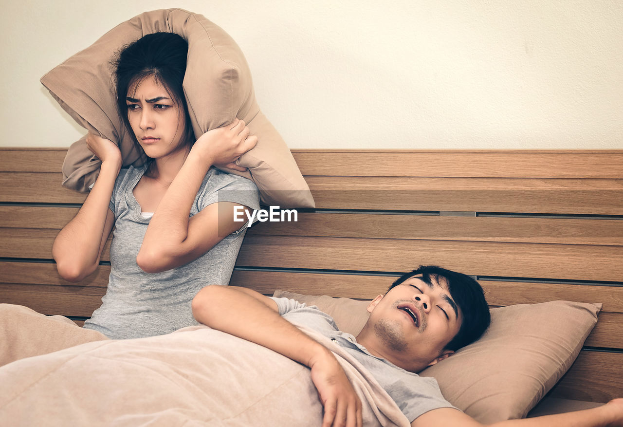 Woman covering ears with cushion while sitting by man sleeping on bed at home