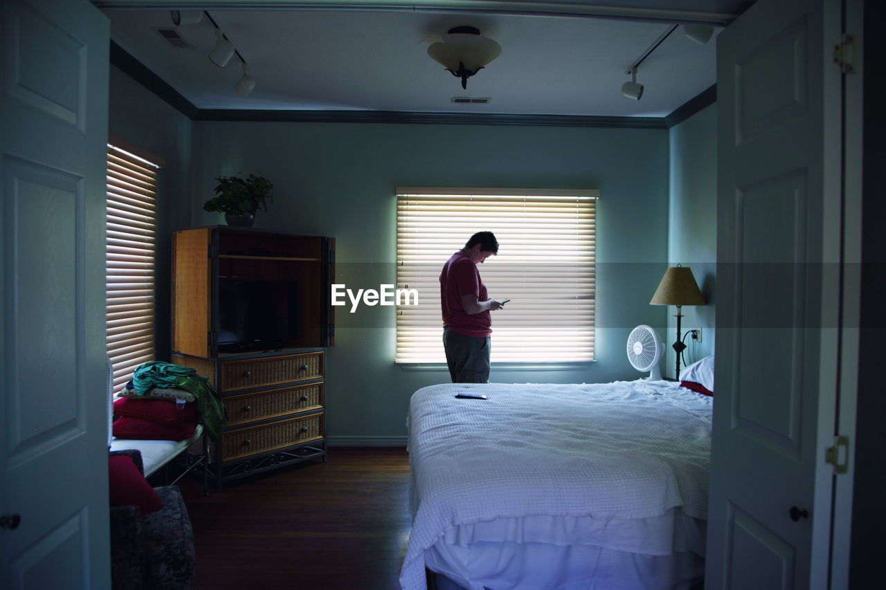 Woman using smart phone while standing against window in bedroom