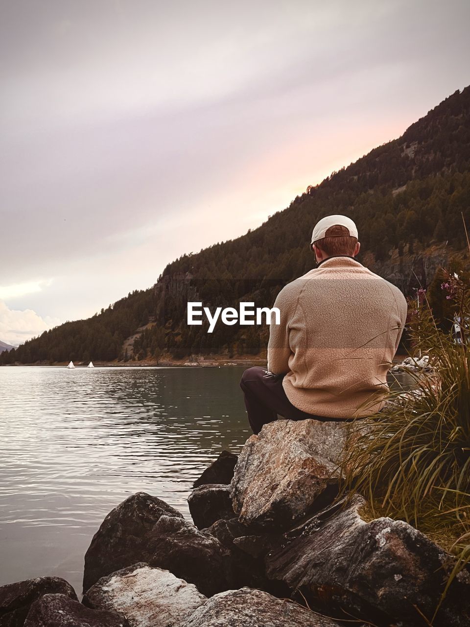 Rear view of man sitting on rock against sky by lake