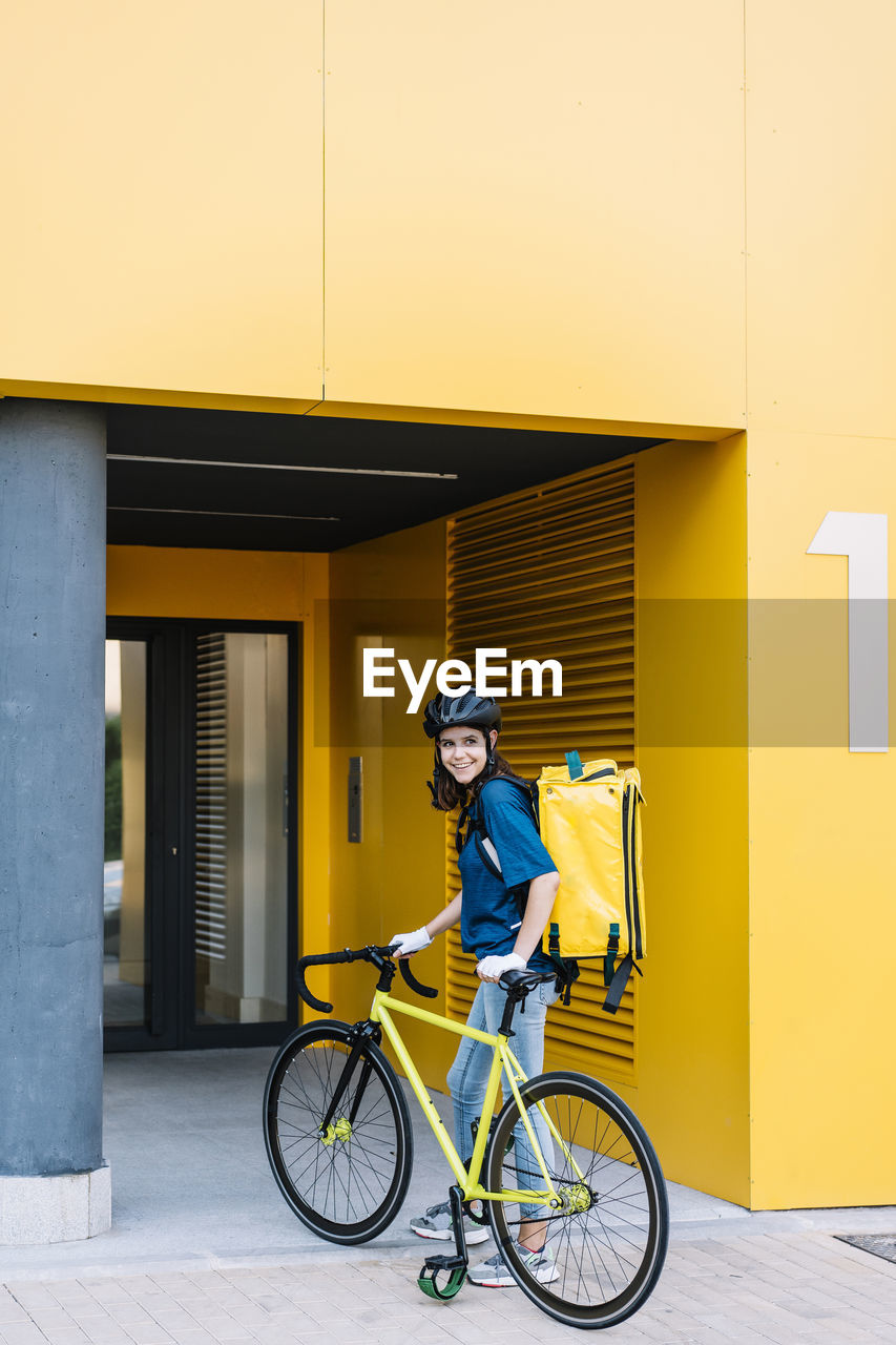 Smiling young delivery woman with bicycle standing in front of building