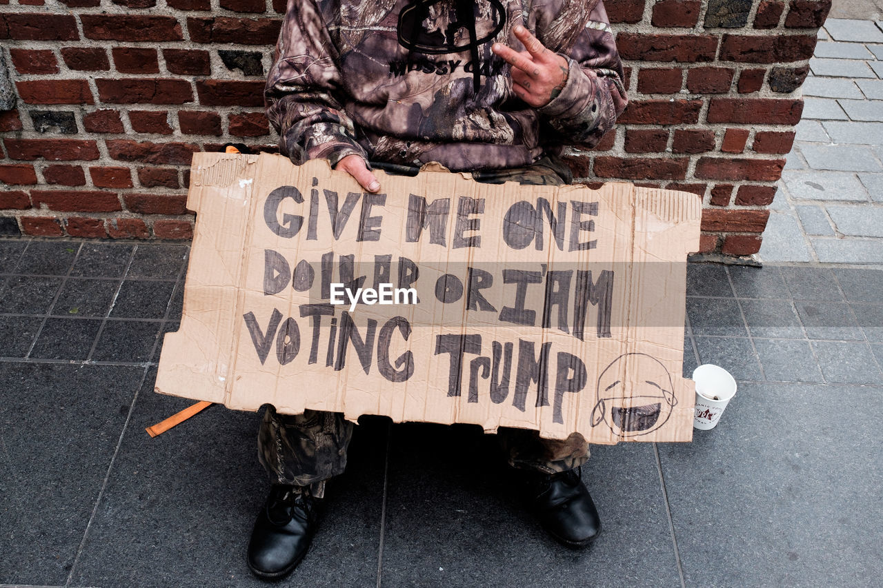 Low section of beggar with sign sitting by brick wall