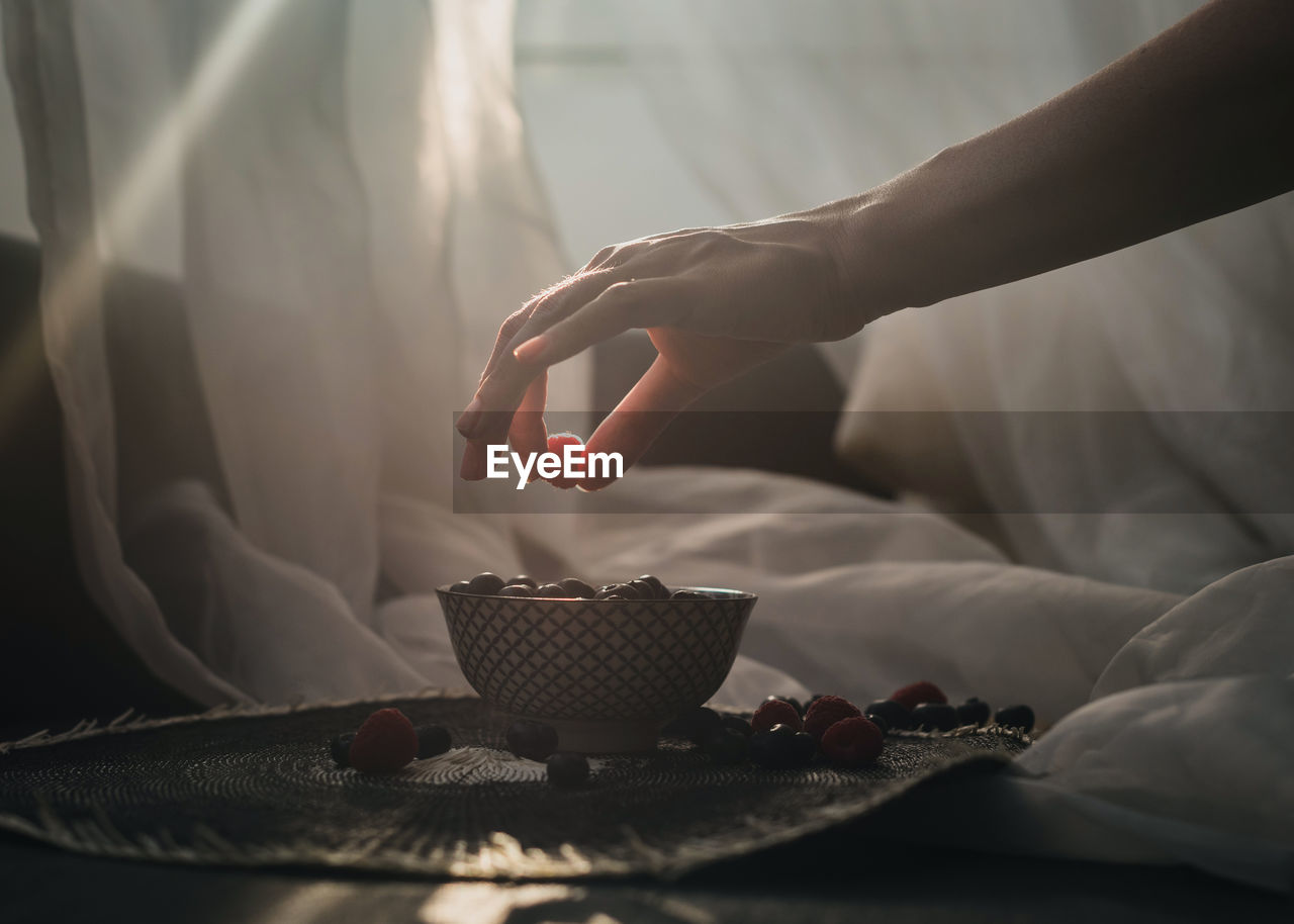 A woman's hand holding a raspberry. underneath is a little bowl more blueberries.