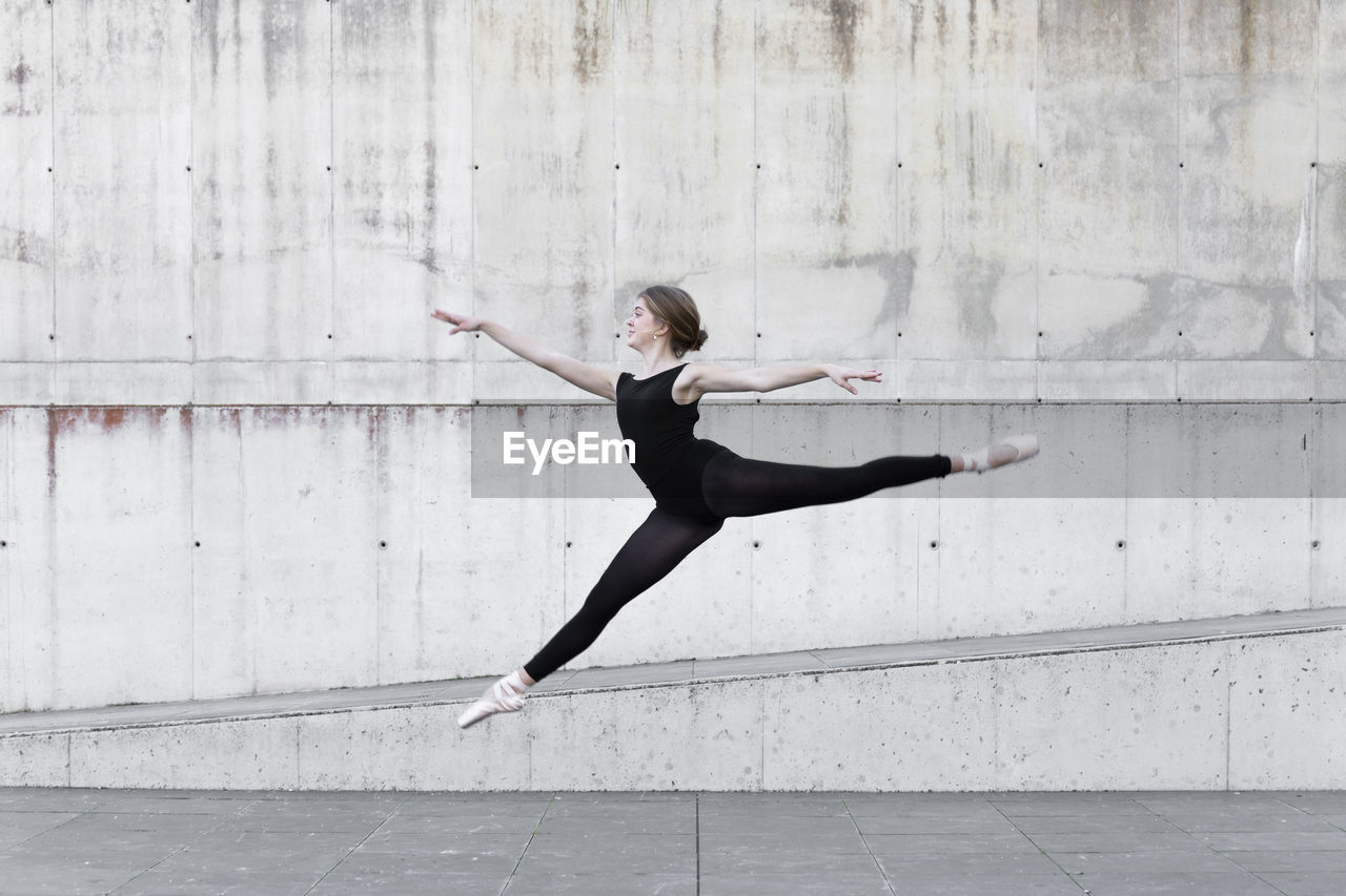 Ballerina in black leotard jumping in front of concrete wall