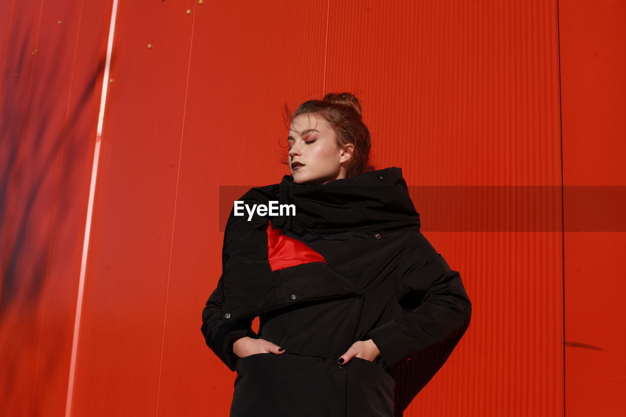 Fashionable teenage girl wearing black warm clothing standing against red wall during sunny day