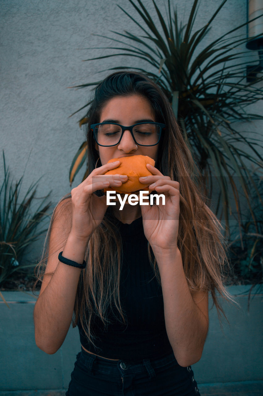 Young woman eating burger while standing against wall