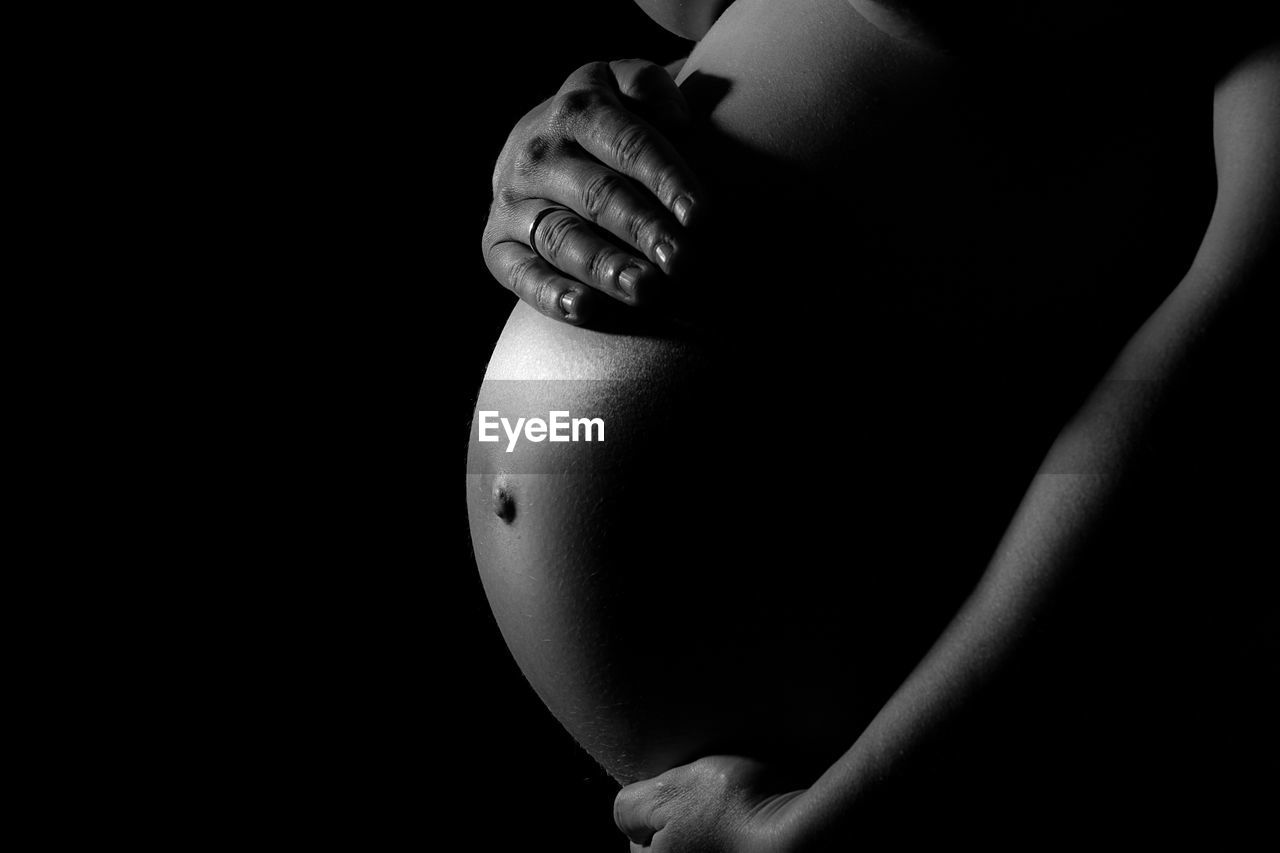 Midsection of pregnant woman touching belly against black background