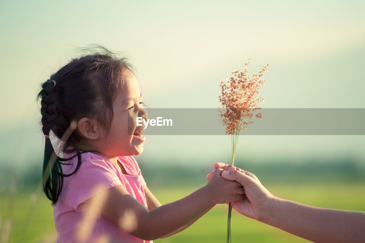 Cropped hand of mother giving flowers to happy daughter against sky