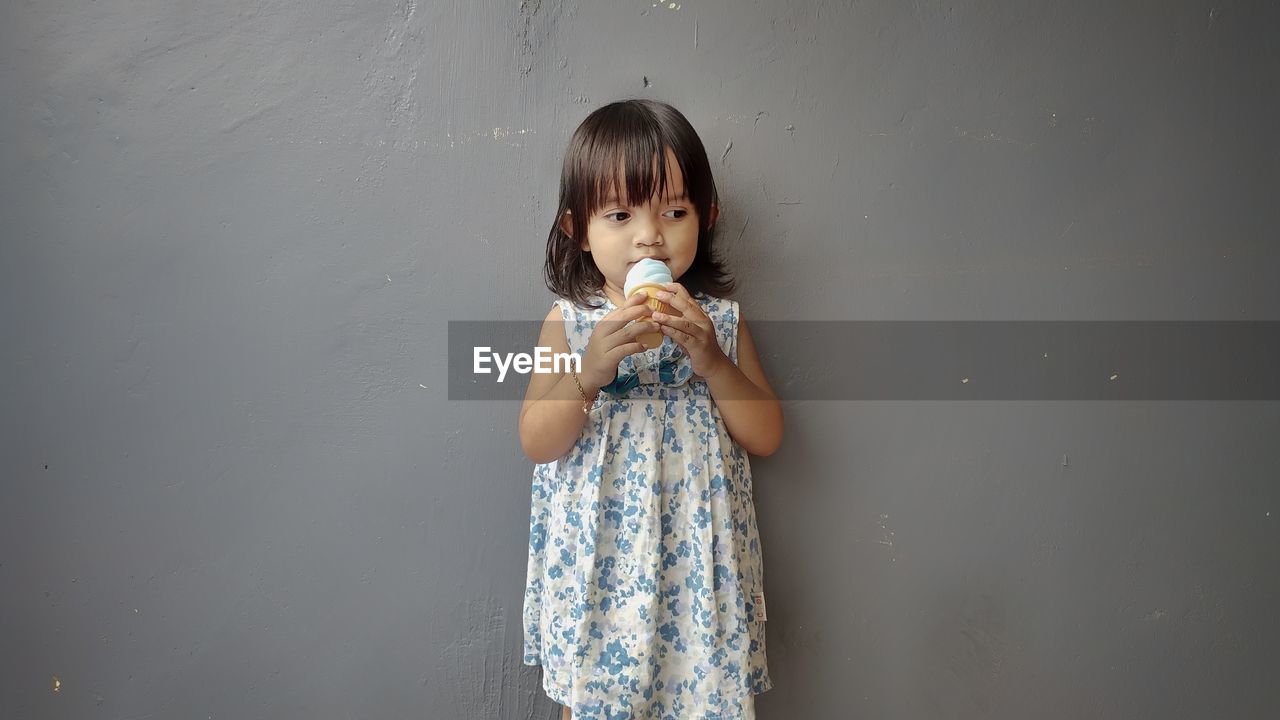Girl eating ice cream while standing against gray wall