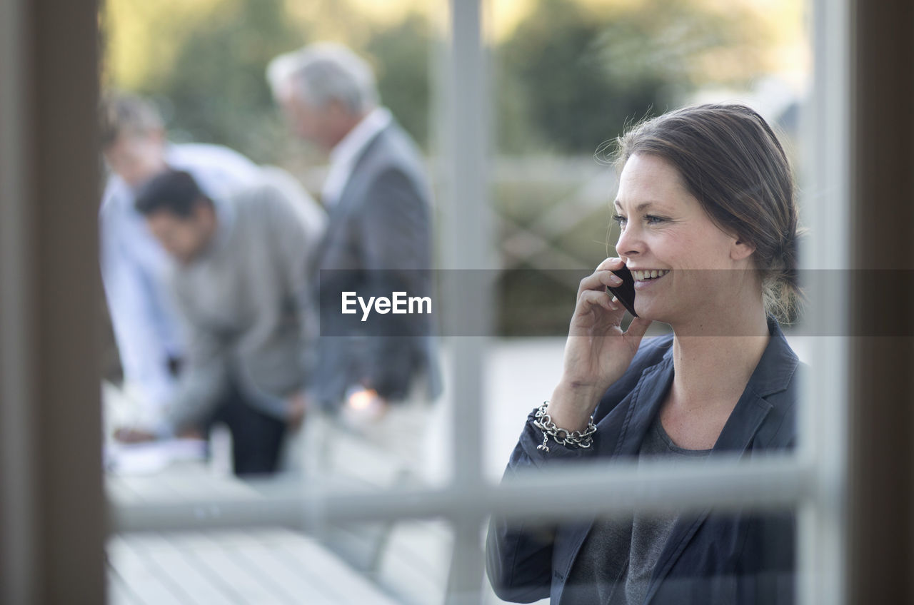 Businesswoman using mobile phone at patio with colleagues working in background