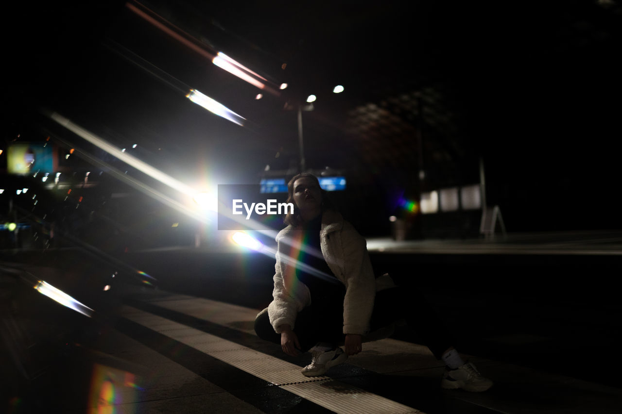 Woman crouching on street against illuminated lights at night