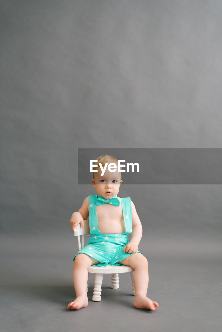 A one-year-old boy is sitting on a white children's chair on a gray background. copy space