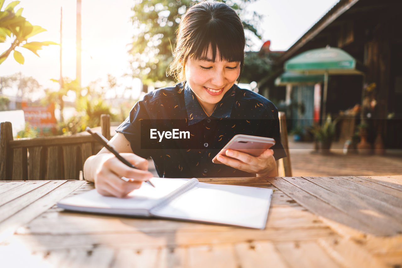 Woman using mobile phone while writing in book on table