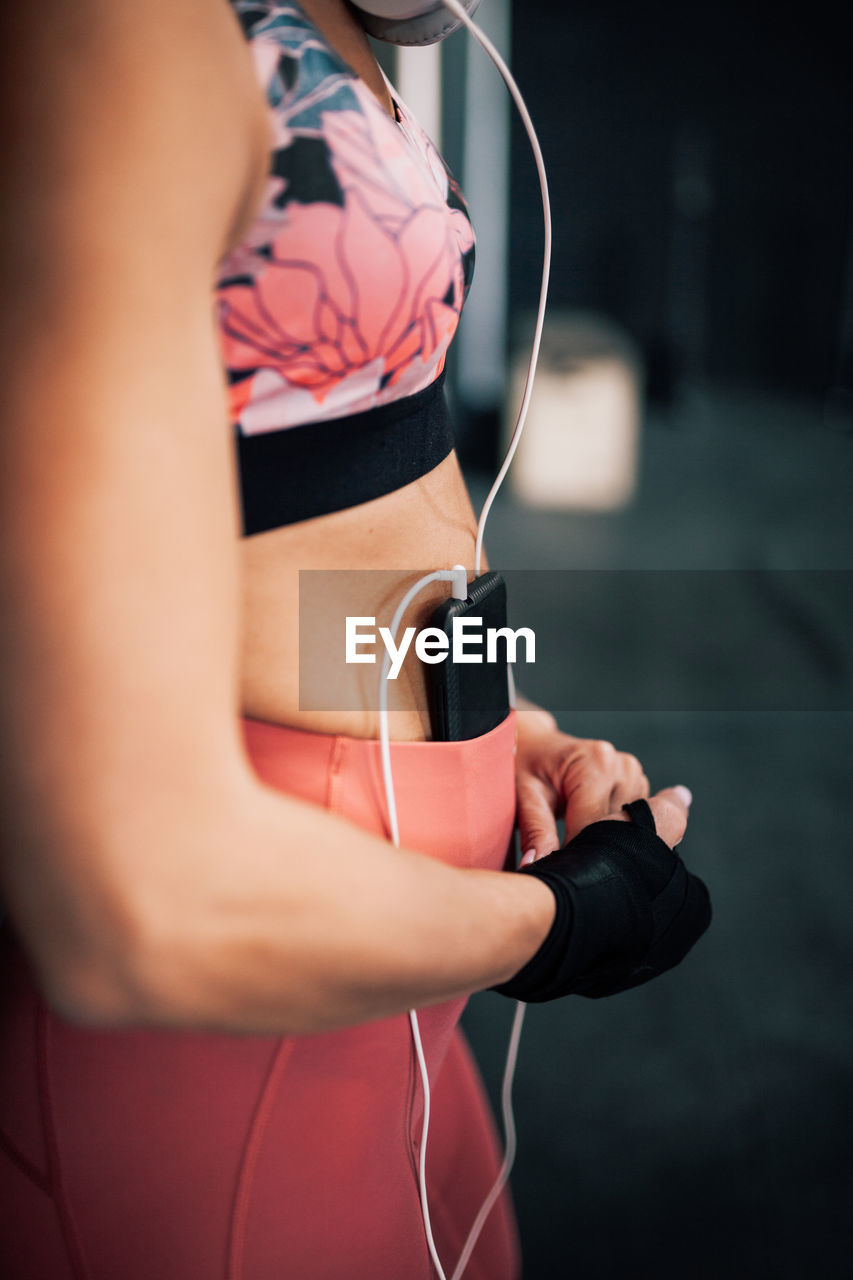 Side view of crop female boxer in sportswear with bandage on wrist listening to music with smartphone and headphones while resting after training in gym