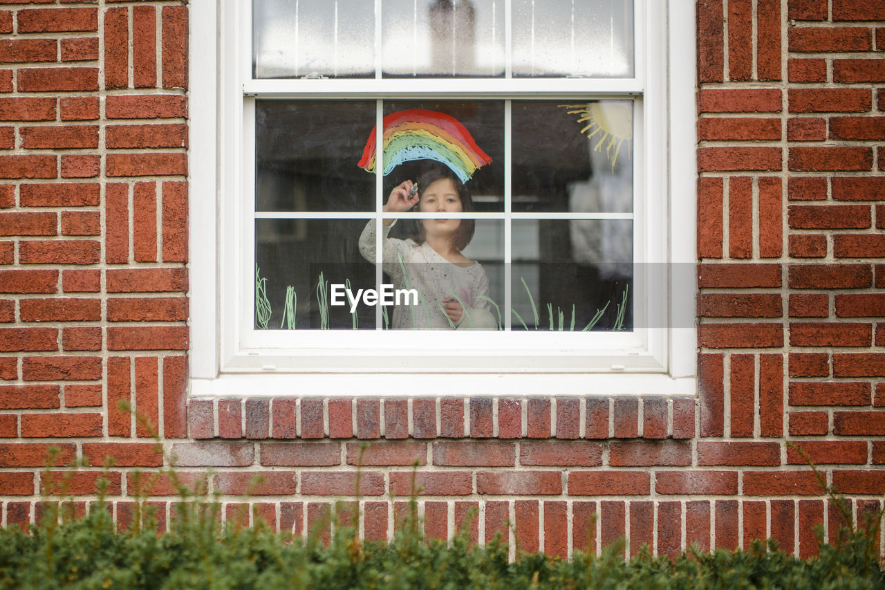 View from outside of small child standing in window drawing a rainbow