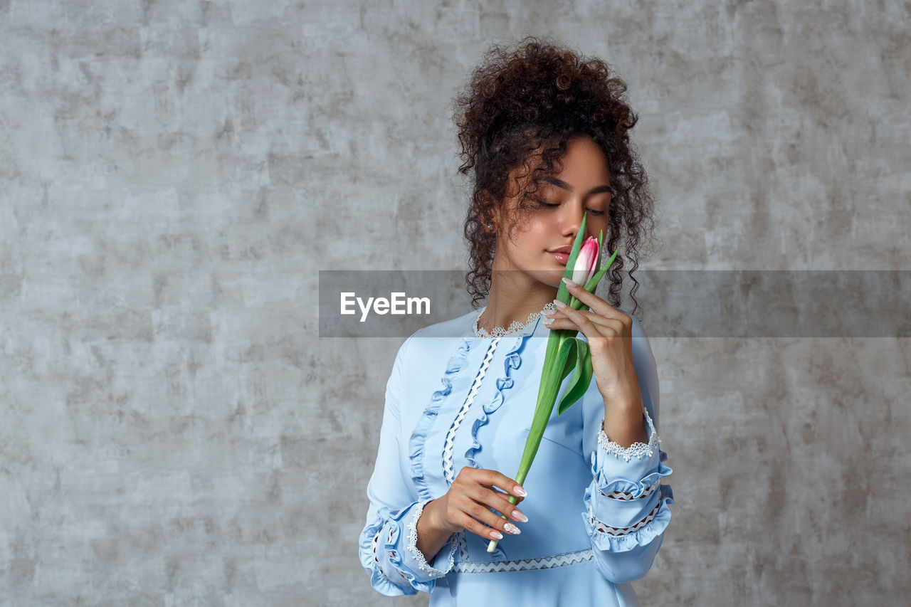 Young woman holding tulip while standing against wall