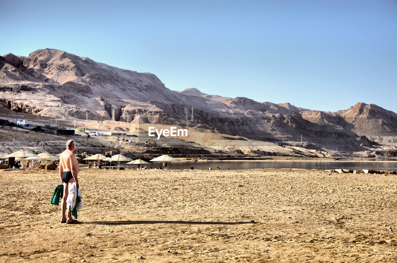 Full length of man standing at beach by rocky mountains against clear sky